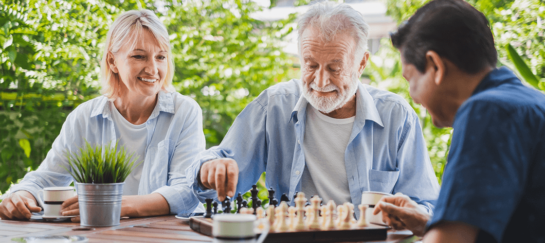 Three elderly people playing chess together outside to improve their mental acuity